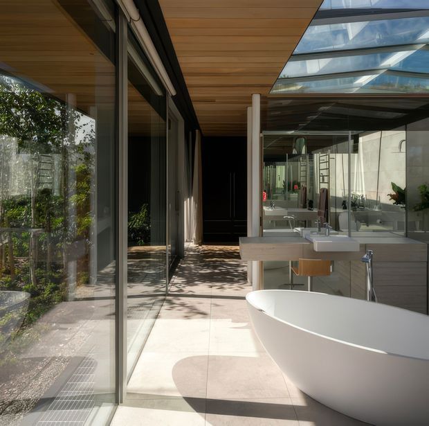 A bright, contemporary bathroom with a freestanding white oval bathtub in the foreground, a floating double vanity with integrated basins and mirrors, large-format pale floor tiles, extensive glazing, and a warm timber-lined ceiling. The bathroom opens onto a planted outdoor area through full-height glass, with a glazed roof providing abundant natural light.