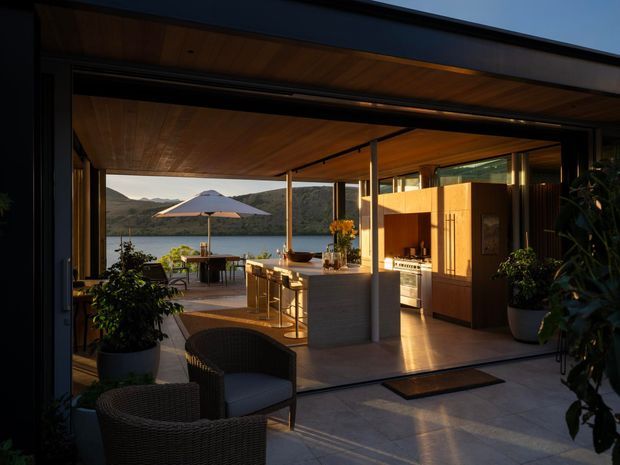 A warm, open-plan kitchen and outdoor entertaining area viewed from a covered terrace. The space features extensive glazing, timber-lined ceilings, dark-framed openings, a pale stone kitchen island with bar seating, timber cabinetry, and a built-in oven. Wicker lounge chairs and potted plants occupy the foreground, while a dining setting and umbrella overlook a calm waterfront landscape beyond.