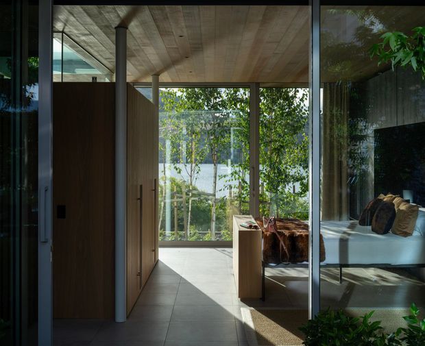 A refined modern interior viewed through large dark-framed glass doors, showing a bedroom beyond a tiled passage. The space features extensive floor-to-ceiling glazing, warm timber wall and ceiling surfaces, slim structural columns, built-in timber joinery, and strong visual connection to leafy landscaping outside. The bedroom contains a low bed with neutral bedding and a dark upholstered throw or cushion, while sunlight creates geometric shadows across the floor.