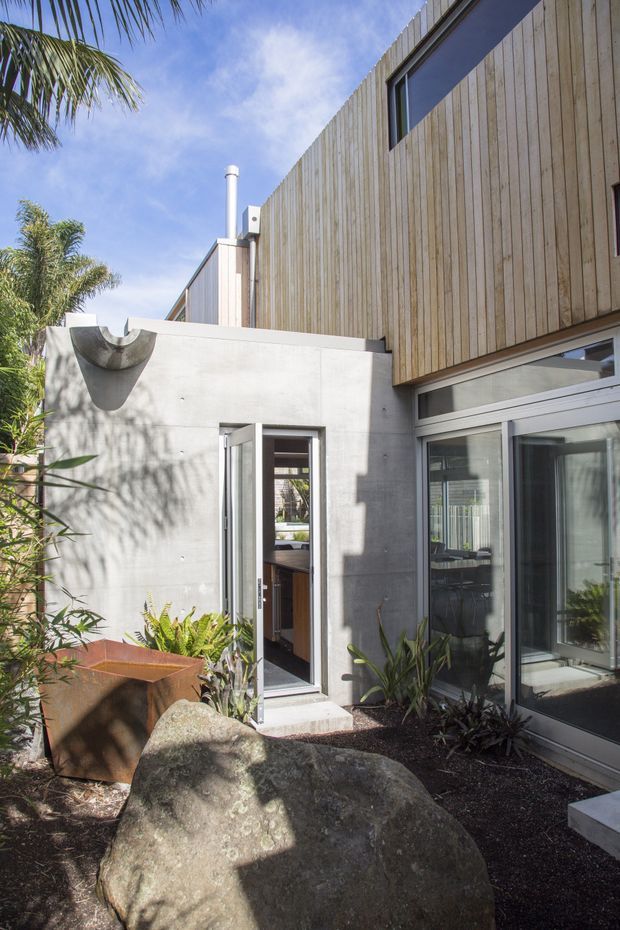 A sunlit modern residential courtyard with a raw concrete wing, vertical timber-clad upper level, and extensive glazed doors and windows. Tropical planting, dark gravel, a large natural boulder, and a rusted metal planter create a landscaped entry garden. The composition highlights the building exterior and its connection to the courtyard.