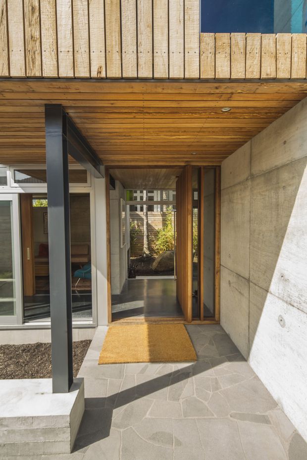 A modern residential entryway viewed from outside, with a covered porch and open timber-lined soffit. The entry combines raw concrete walls, dark metal structural posts, timber cladding and door frames, large glazed openings, and irregular grey stone paving. A warm-toned doormat leads into a glazed hallway, with a small landscaped courtyard visible beyond.