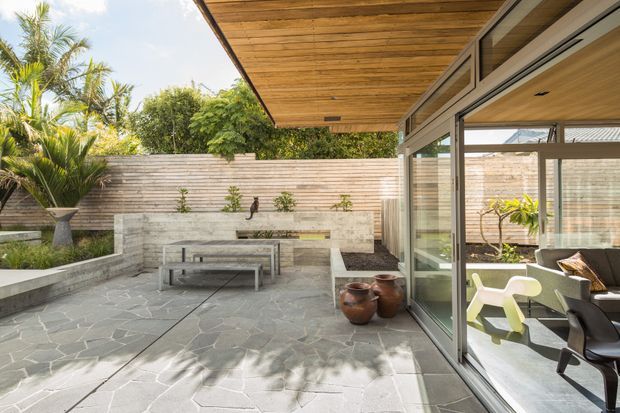 A bright, modern indoor-outdoor entertaining area opening onto a landscaped courtyard. The terrace has irregular grey stone paving, a built-in rendered bench and raised planter, a horizontal timber privacy screen, and a broad timber-lined roof overhang. Full-height glazed sliding doors connect the terrace to a dining area with contemporary seating and timber ceilings. Tropical planting and strong indoor-outdoor continuity create a relaxed, resort-like setting.