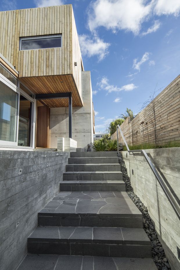 Exterior view looking up a narrow stepped walkway toward a modern multi-level home. The facade combines vertically clad light timber, exposed concrete walls and soffits, large glazed openings with dark frames, and dark stone paving. Concrete retaining walls, timber privacy fencing, planting, and a metal handrail define the sloped entry approach under a bright blue sky.