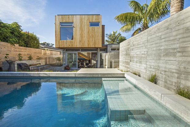 Contemporary multi-storey residence with warm vertical timber cladding, extensive glazing and a sculptural concrete pool terrace. The foreground features a swimming pool with pale tiled shallow steps and submerged seating, bordered by a tall raw concrete wall and timber privacy fencing. Tropical palms and planting surround the outdoor entertaining area.