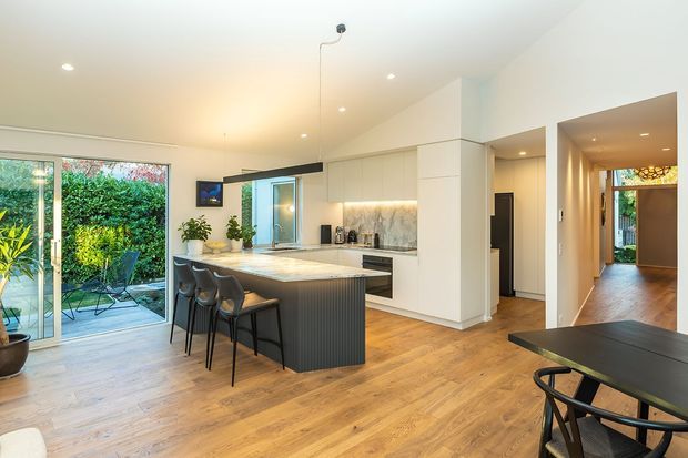Bright open-plan kitchen and dining area with a central island and seating, white built-in cabinetry, stone-look splashback, timber flooring, and large sliding glass doors opening to a leafy outdoor area. A hallway and additional rooms are visible beyond the kitchen.