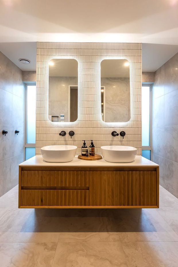 A refined contemporary bathroom centred on a floating double vanity with two vessel basins, dark wall-mounted tapware, and integrated drawer cabinetry in warm fluted timber. The vanity has a pale stone benchtop and sits against a vertical ribbed feature wall with two rounded-rectangle backlit mirrors. Large-format pale stone-look tiles cover the walls and floor, while narrow windows on either side provide natural light. The overall palette is warm, neutral, and softly illuminated.