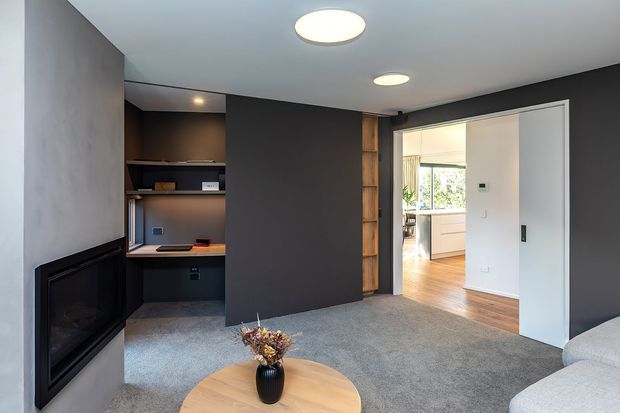 A contemporary living room with dark charcoal feature walls, grey carpet, recessed circular ceiling lights, a built-in black fireplace, and a compact integrated study nook with shelving and desk. A timber-framed vertical shelving detail and large white sliding door open toward a bright kitchen or dining area with timber flooring. The edge of a light sofa and round timber coffee table are visible.