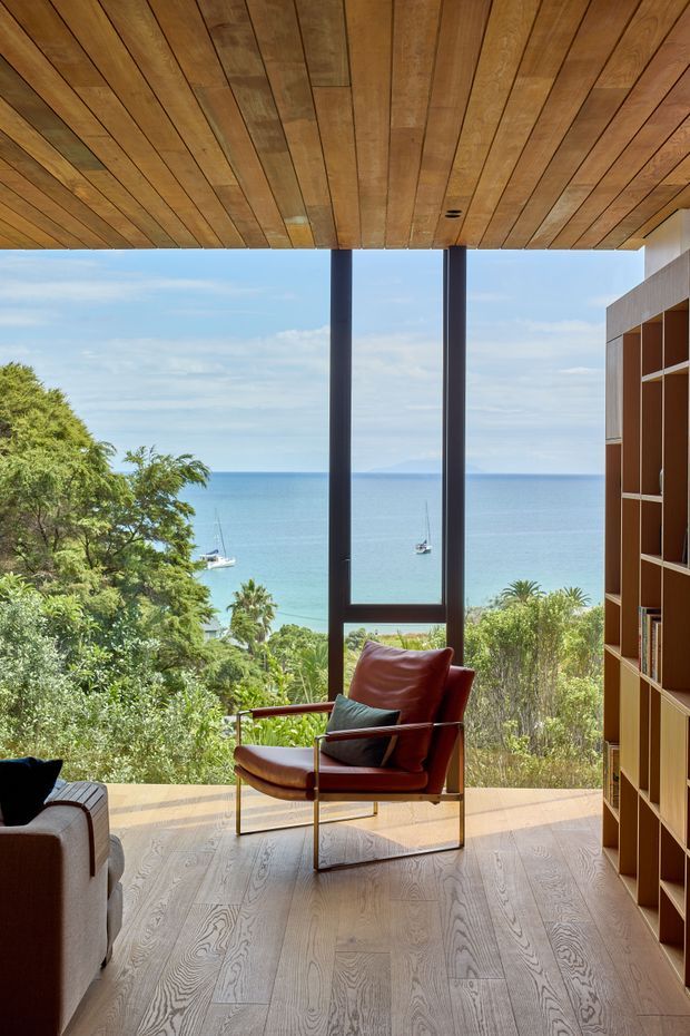 A serene coastal living room with expansive floor-to-ceiling glazing overlooking a blue ocean, boats, tropical vegetation, and a distant shoreline. The room features a warm timber-planked ceiling, pale timber flooring, slim dark-framed glazing, a built-in timber shelving wall, and a sculptural leather lounge chair. A partial sofa is visible in the foreground. The composition emphasises the indoor-outdoor connection and panoramic coastal outlook.