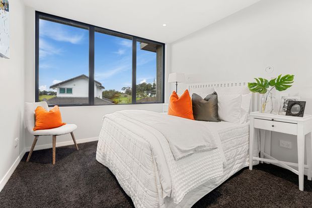 Bright contemporary bedroom with a white upholstered bed dressed in textured white bedding, vivid orange and dark grey accent cushions, a white bedside table, and a small white chair. Large black-framed windows provide expansive suburban views and abundant natural light. The room has dark charcoal carpet, white walls, and a clean, relaxed aesthetic.