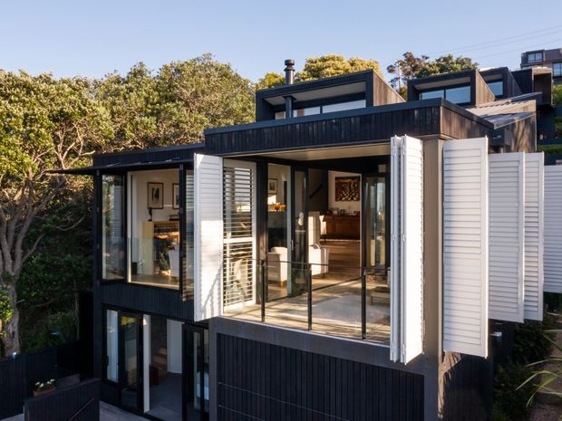 Exterior view of a contemporary multi-level residential building set among mature trees. The facade combines dark vertical cladding with white horizontal weatherboard panels, large glazed openings, projecting box-like volumes, and a terrace or balcony with glass balustrading. White external louvred shutters are opened beside the upper-level glazing, and a dark roofline with chimney elements is visible.