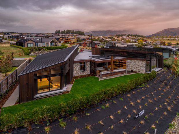 Aerial exterior view of a contemporary single-storey residence with dark metal pitched roofs, timber and dark cladding, extensive glazing, and a stone-clad central pavilion. The home surrounds a landscaped lawn and covered outdoor entertaining area, with a suburban neighbourhood and mountainous backdrop beyond.