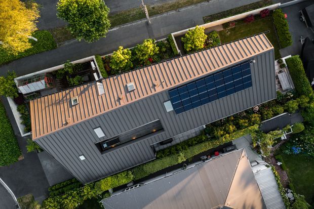 Aerial top-down view of a long contemporary residential building with intersecting metal-clad pitched roof forms, multiple skylights, and a large array of dark solar panels. The property is surrounded by established landscaped gardens, paved paths, and neighbouring residential structures.