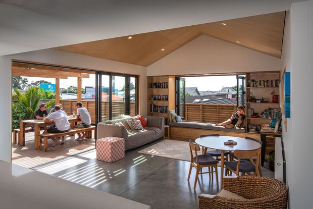 A bright, open-plan living and dining space with polished concrete flooring, extensive timber-framed glazing, built-in timber shelving, and a dramatic pitched timber ceiling feature. The room opens onto a timber deck where people are gathered around an outdoor dining table, with a sofa, round dining table, and woven furniture inside. Strong natural light creates a relaxed indoor-outdoor connection.