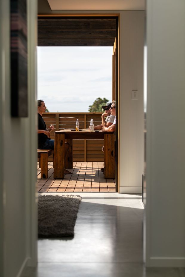 A view from inside a minimalist, light-filled interior through an open doorway onto a timber deck. The deck features a substantial rustic timber dining table with bench seating, where several people are gathered. Horizontal timber screening and warm wood framing surround the outdoor opening, while the foreground has pale smooth flooring and a textured dark rug.
