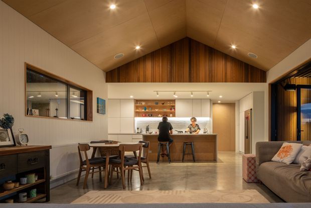 Warm, open-plan contemporary living and dining area with a vaulted timber-lined ceiling. The room combines a timber dining table and chairs, a compact kitchen with a central island and stools, built-in cabinetry, polished concrete flooring, and a comfortable sofa seating area. Vertical timber panelling forms a feature wall beneath the gable, while integrated downlights create a welcoming evening ambience.