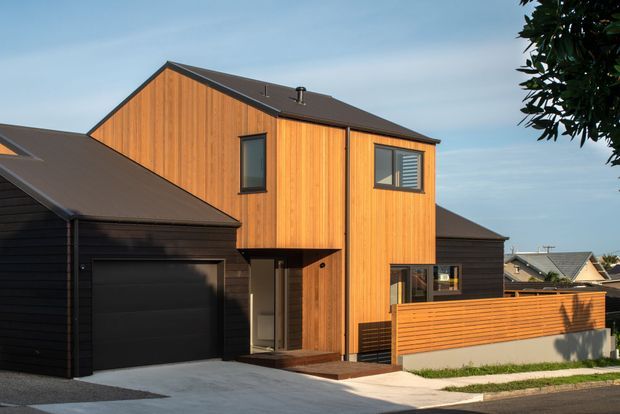 Exterior view of a contemporary multi-level residence with warm vertical timber cladding, a dark pitched roof, integrated garage, recessed entry, and a horizontal timber privacy fence. The home sits in a compact suburban streetscape with neighbouring houses visible in the background.