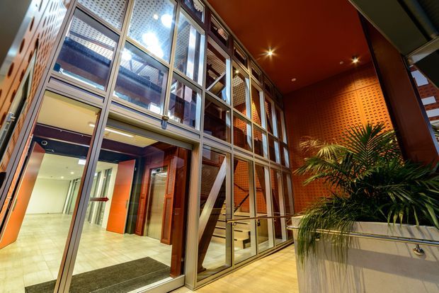 A dramatic multi-level commercial or institutional interior atrium with a tall glazed curtain wall, exposed stair, warm orange-red ceiling and feature brick wall, perforated metal screening, tiled flooring, and a large indoor palm planter. The entry lobby and circulation space are brightly lit with strong architectural glazing and warm materials.