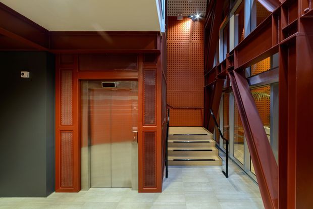 Interior view of a compact contemporary stairway and elevator lobby. The space features a polished light-grey floor, a stainless-steel elevator set within reddish-brown timber or metal wall panelling, perforated acoustic panels, black handrails, and exposed structural bracing alongside tall glazed openings. A short flight of stairs leads upward through the circulation space.