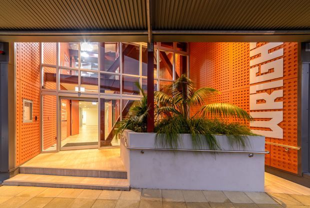 A covered commercial entry courtyard or lobby with a bold orange perforated metal wall treatment, exposed dark steel framing, extensive glazed doors and windows, and a ribbed metal ceiling. A large raised concrete planter with tropical palms sits in the foreground, while illuminated interior circulation spaces are visible beyond the glass. Large white vertical lettering forms a prominent feature on the right wall.
