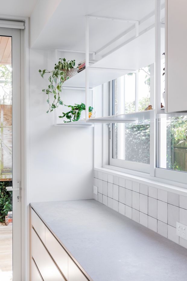 Bright, narrow kitchen workspace with a long pale grey benchtop, white drawer cabinetry, tiled splashback, large white-framed windows, and open white shelving. Hanging and potted greenery adds a natural accent, while the predominantly white fit-out creates an airy, minimalist appearance.