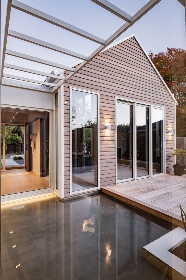 Contemporary courtyard exterior showing a small gabled wing clad in horizontal timber weatherboards, tall glazed doors and windows, and a timber deck beside a shallow reflective water feature. A white-framed glazed pergola or covered walkway connects to the adjoining interior, with warm interior lighting and landscaped planting visible around the edges.