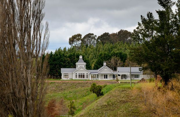 A long, single-storey country residence set within a landscaped rural property. The light-coloured building has a symmetrical gabled central section, classical decorative timber detailing, a prominent chimney, extensive windows, and low pitched roof wings. Tall poplars, mature trees, open grass, and gently sloping grounds surround the house under an overcast sky.