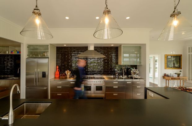 A spacious contemporary kitchen viewed from behind a large dark island with an integrated sink and curved stainless-steel tap. The room features extensive dark wood cabinetry, stainless-steel appliances, a dark glossy brick-tile splashback, glass-fronted upper cabinets, a large central range cooker with a stainless-steel canopy hood, and three clear pendant lights. White walls and ceiling provide contrast, while a doorway and dining or service area are visible to the right.