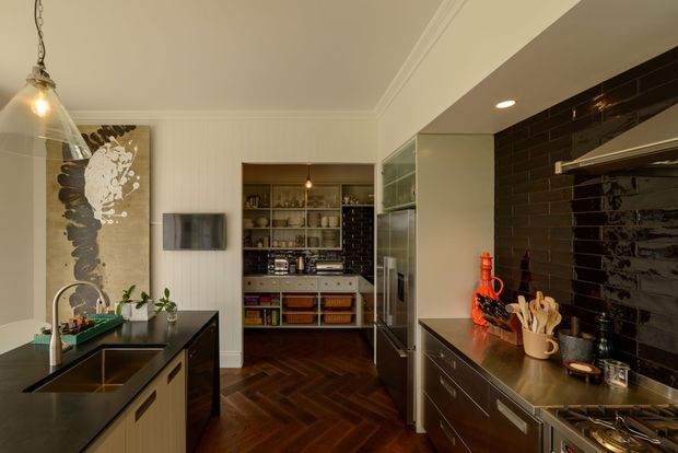 A warm, refined contemporary kitchen viewed toward a compact butler's pantry or scullery. The main kitchen has dark benchtops, dark glossy subway-tile splashbacks, extensive dark cabinetry, a contrasting light island with an integrated sink, stainless-steel appliances, and a rich dark timber herringbone floor. White vertical wall panelling, glass-fronted storage, pendant lighting, and open pantry shelving complete the space.