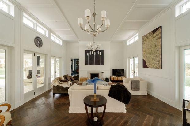 Bright, refined living room with white paneled walls, a coffered or panelled ceiling, tall clerestory windows, and extensive glazed doors. The room features a herringbone dark timber floor, a central fireplace, upholstered seating, artwork, and elegant multi-arm chandeliers. The overall design combines traditional architectural detailing with contemporary furnishings and a light, airy atmosphere.