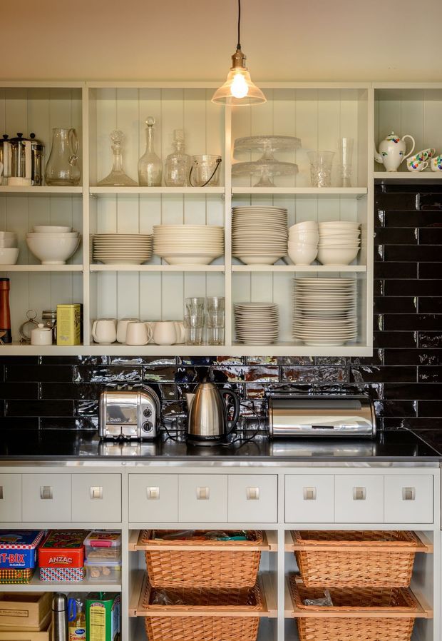 A close-up view of a well-organised kitchen pantry or storage wall. White open shelving displays stacked plates, bowls, glassware, cake stands, and decorative crockery, while lower drawers and pull-out wicker baskets provide additional storage. The cabinetry and shelving are painted off-white, with a glossy black tiled backsplash and dark benchtop. A pendant light hangs above the shelving, and stainless-steel countertop appliances sit below.