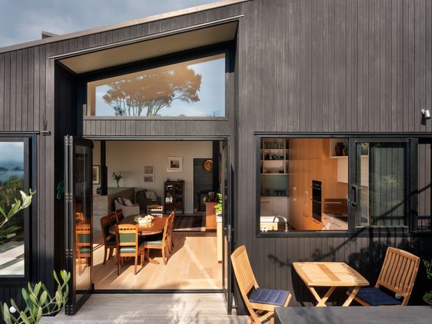 Exterior view of a contemporary dark-clad home opening onto a timber deck or patio. Large black-framed glazing and folding or sliding doors reveal a dining and living area inside, while a kitchen with warm timber cabinetry is visible through the right-hand window. Outdoor timber dining furniture occupies the foreground, and the upper-level window reflects a tree and sky.