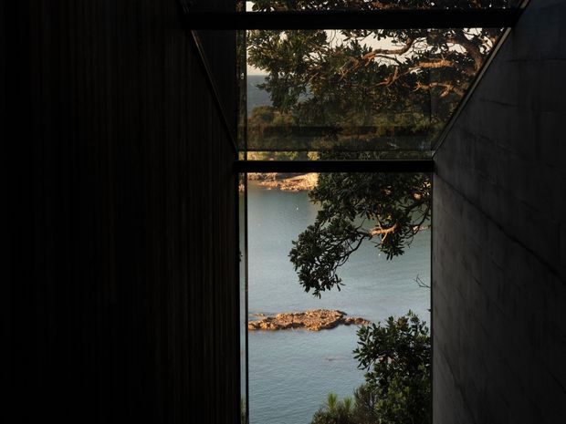 A narrow architectural passage or recessed viewing corridor framed by dark vertical cladding on the left and a dark solid wall on the right. Large glazed openings at the end and overhead create a carefully framed view toward calm blue water, rocky shoreline, and surrounding coastal vegetation. The scene is dim, dramatic, and strongly focused on the relationship between the built structure and the landscape.