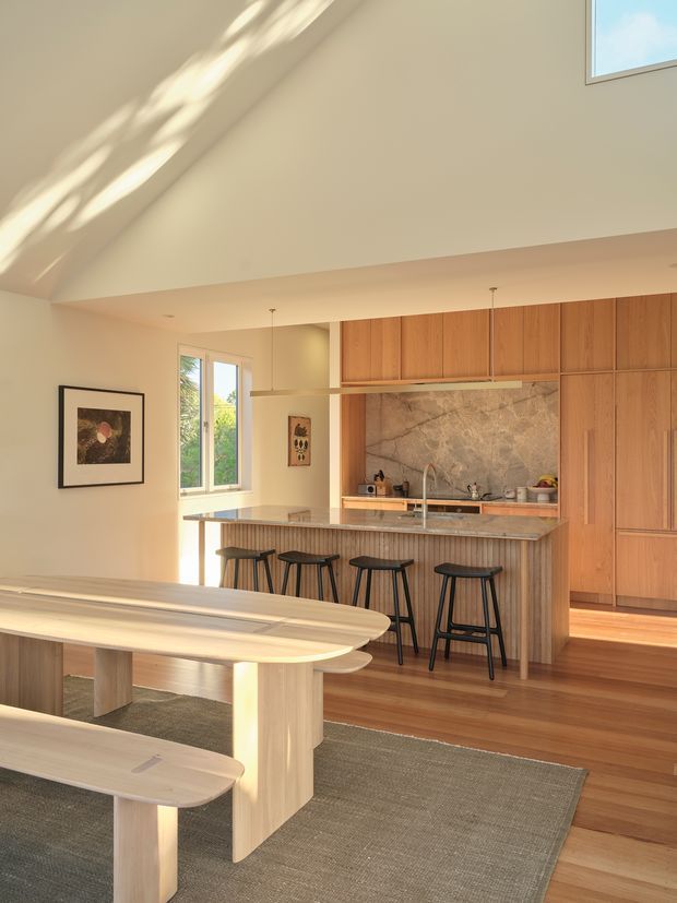 Warm, light-filled open-plan kitchen and dining area with a high sloping ceiling and clerestory-style glazing. The kitchen features extensive timber cabinetry, a large stone island with integrated seating, a textured stone splashback, and timber flooring. A sculptural timber dining table and benches occupy the foreground, creating a refined contemporary interior.