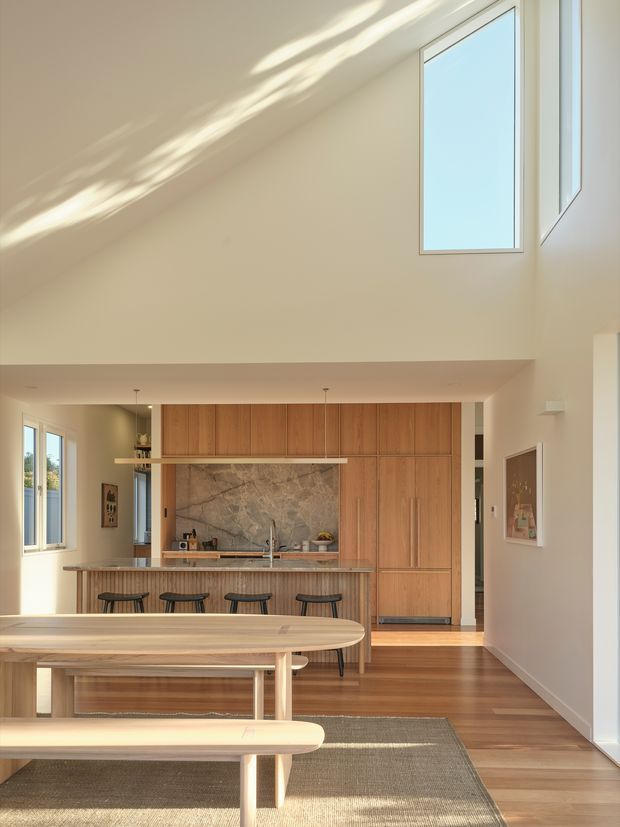 Bright, double-height open-plan kitchen and dining area with warm timber cabinetry, a large stone feature splashback, island breakfast bar with stools, and a long timber dining table with benches. Tall windows and clerestory glazing fill the space with sunlight, while polished timber flooring and neutral walls create a warm, contemporary interior.