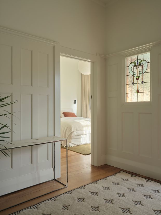 A bright, refined residential entryway with white paneled walls and doors, detailed cornices, a timber floor, and a large patterned rug. A slim console table sits against the panelled wall, while a stained-glass front door provides a decorative focal point. Through the open doorway, a softly styled bedroom with a bed, curtains, and timber flooring is visible.