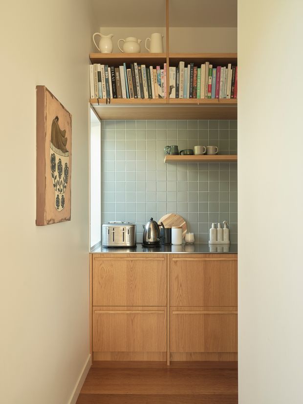 Compact built-in kitchen or kitchenette viewed from a hallway, featuring warm timber cabinetry, a dark benchtop, muted green square-tile splashback, open shelving with books and ceramics, and countertop appliances. The narrow space has a warm, curated mid-century-inspired character.