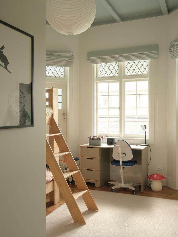 Bright, calm children's bedroom with a built-in study nook beneath tall leadlight-style diamond-pane windows. The room features a timber loft bed and ladder, pale desk and drawers, child-sized swivel chair, carpeted floor, white walls, roller blinds, exposed ceiling beams, a large paper pendant light, and playful mushroom-shaped decor.