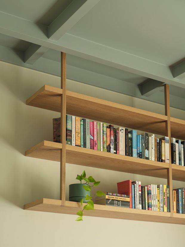 Close-up interior view of a built-in open timber shelving system spanning a wall, filled with books and a small potted plant. The shelves are supported by slim vertical timber posts and sit beneath a pale painted ceiling with exposed structural beams. The palette is warm wood, muted green, and soft neutral tones.