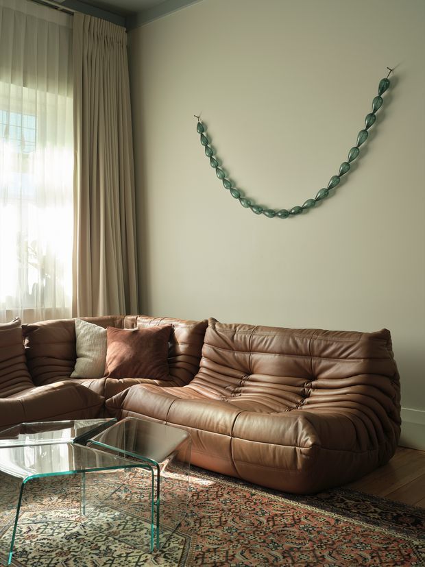 A warmly lit living room with a low, tufted brown leather sectional sofa, patterned area rug, and transparent glass coffee table. Full-height curtains frame a bright window, while a simple green botanical wall decoration adds a subtle feature against the pale neutral wall. Timber flooring and layered natural textures create a relaxed, inviting atmosphere.