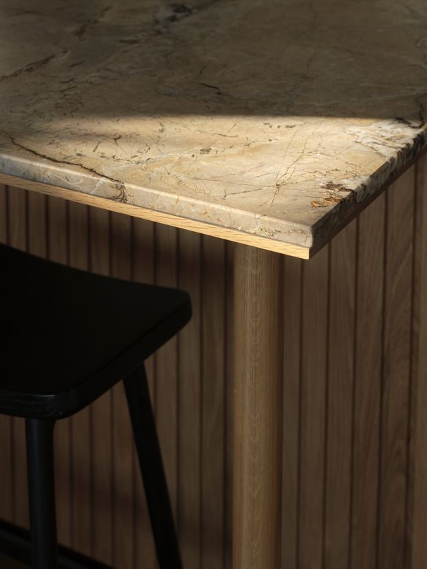 Close-up of a small bar-height table or counter with a thick, irregularly edged beige veined stone top, supported by a round timber leg. Vertical dark timber wall panelling forms the backdrop, with a black stool partially visible at left. The image has warm, moody lighting and focuses on the material and joinery detail.