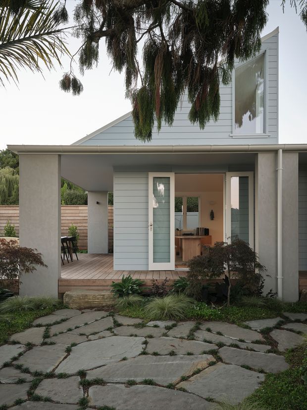 Front exterior view of a light grey weatherboard house with a covered entry porch, open glass door, pale timber decking, simple vertical columns, and a landscaped garden. Irregular large stone paving forms the foreground path, while mature trees partially frame the facade. The image also reveals a glimpse of the kitchen and dining area through the open doorway.