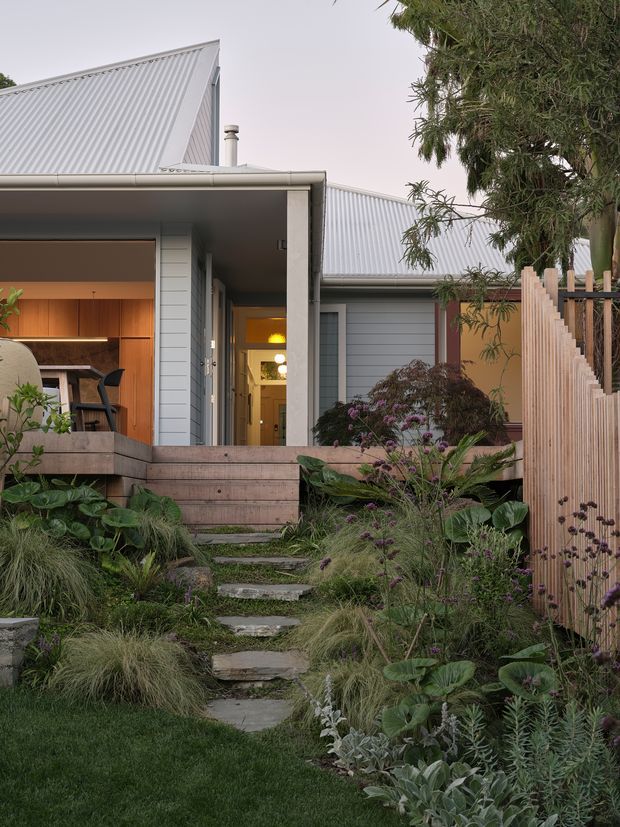 Exterior view of a contemporary single-storey house with light grey weatherboard cladding, corrugated metal gable roofs, and a covered entry terrace. A timber-lined interior and warm lighting are visible through the open recessed entrance. The house is approached by irregular stone stepping slabs through a lush, naturalistic garden with ornamental grasses and layered planting, alongside a vertical timber privacy fence.
