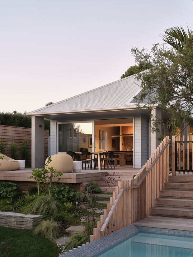 Exterior view of a contemporary single-storey house with a light grey pitched metal roof, pale weatherboard cladding, and extensive open glazing onto a covered outdoor entertaining deck. Timber steps and balustrades lead down through a landscaped garden to a tiled swimming pool in the foreground. Warm interior lighting reveals a kitchen and dining area inside.