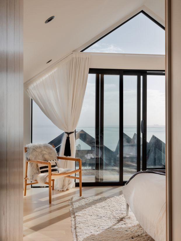 A bright, refined bedroom with expansive black-framed glazing overlooking the ocean and neighboring pitched rooftops. The room features a partially visible bed, pale timber flooring, a cream textured rug, a timber lounge chair with a sheepskin throw, and sheer cream curtains gathered beside the glazing. A dramatic triangular clerestory window and sloping ceiling create an airy architectural focal point.