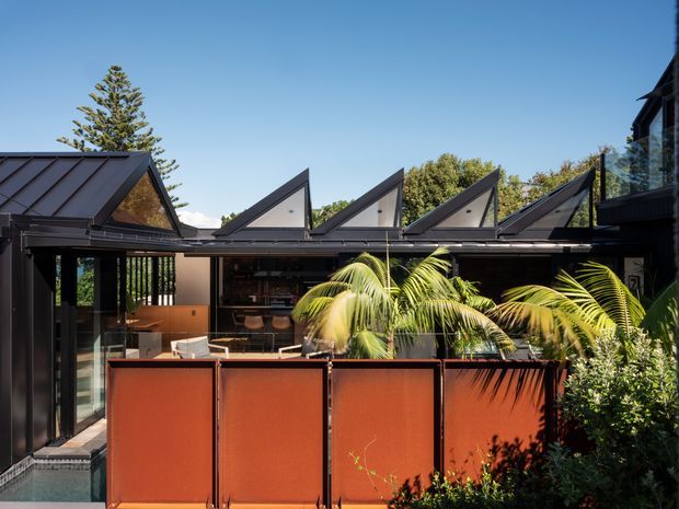 Exterior view of a contemporary residential building with dark metal cladding and a distinctive repeating sawtooth roofline. A glazed outdoor living or dining area opens onto a terrace screened by rust-red privacy panels, with tropical planting and a small visible pool edge in the foreground.