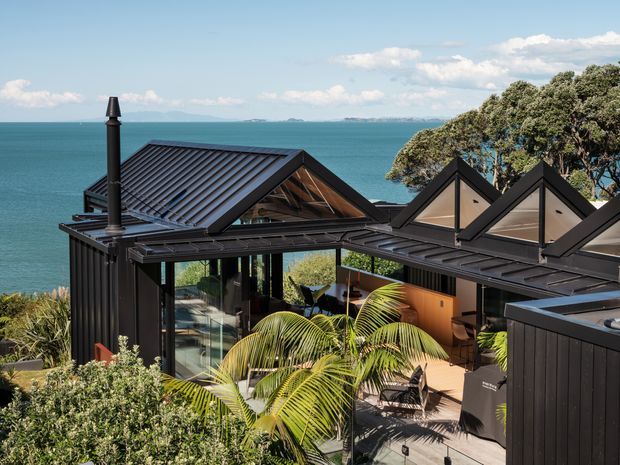 Exterior view of a contemporary dark-clad coastal home with multiple pitched gable roof forms, expansive glazing, and an elevated outdoor deck or terrace. The house overlooks a turquoise body of water, with palms and mature vegetation in the foreground. A dark metal flue is visible on the left roof, and the composition emphasises the building's waterfront setting and angular roofline.