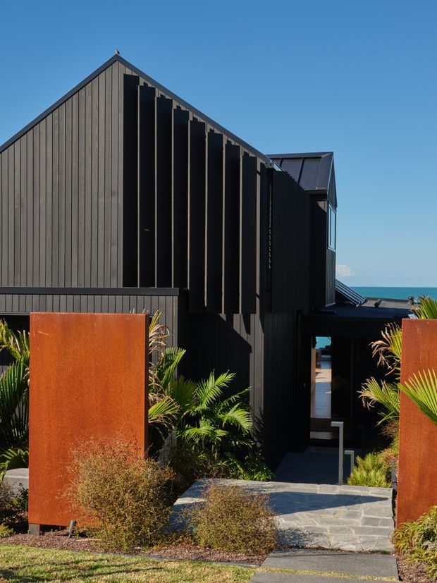 Exterior view of a contemporary dark-clad coastal house, featuring a steep gabled roof, strong vertical timber-like cladding, deep black structural fins, and a recessed entry walkway. Rusted corten-steel garden screens frame lush subtropical planting, with glimpses of blue ocean beyond the building.
