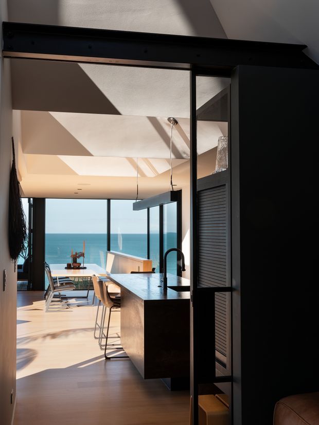 A dramatic view through a dark-framed opening into a bright, contemporary open-plan kitchen and dining area. The space features a large dark kitchen island with an integrated black gooseneck tap, pale timber flooring, a slim linear pendant light, full-height glazing, and a dining table overlooking the ocean. Strong angular sunlight creates geometric shadows across the light ceiling and floor. A dark louvred/slatted fixed screen or cabinetry element frames the right side.