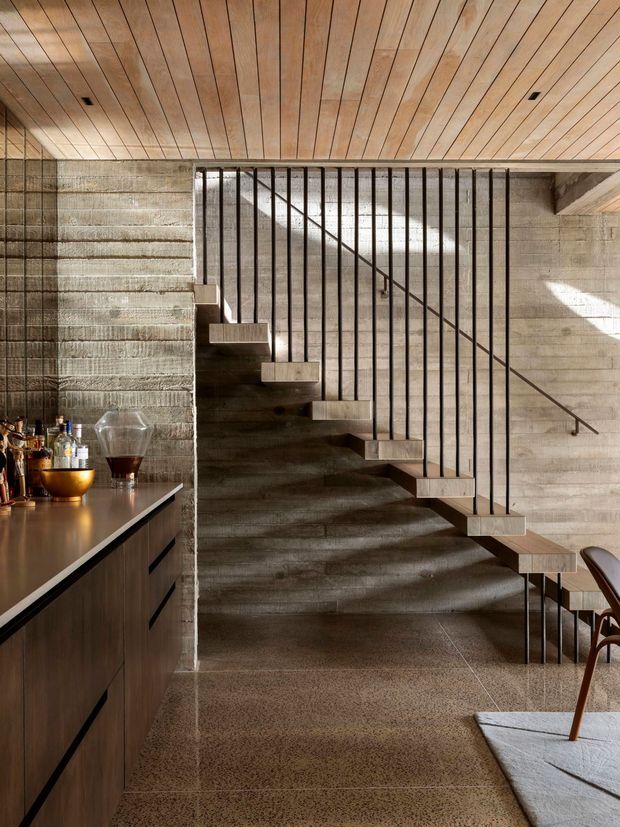Interior view of a refined open-plan space focused on a sculptural concrete staircase with floating treads and slender black vertical balustrades. A dark timber built-in bar or cabinetry run with bottles and glassware occupies the left side, while a dining chair and table edge appear at right. The room features exposed textured concrete walls, a warm timber-lined ceiling, and a polished or honed terrazzo-like concrete floor. Natural light creates strong geometric shadows across the staircase and walls.