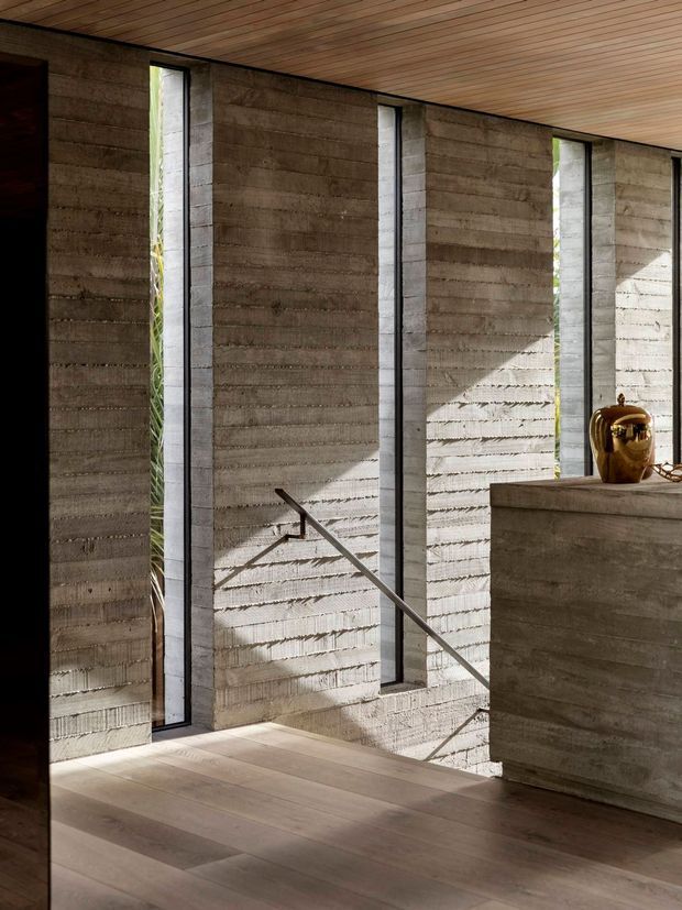 A minimalist interior entry or stair landing with heavily textured board-formed concrete walls, narrow vertical windows, a slim metal stair handrail, and a warm timber slatted ceiling. Wide pale timber flooring and a substantial concrete console or partition occupy the foreground, with strong sunlight casting angular shadows across the wall.